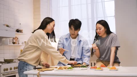 Three Japanese Friends Sitting Around The Kitchen Counter Cutting And Eating Sushi 1