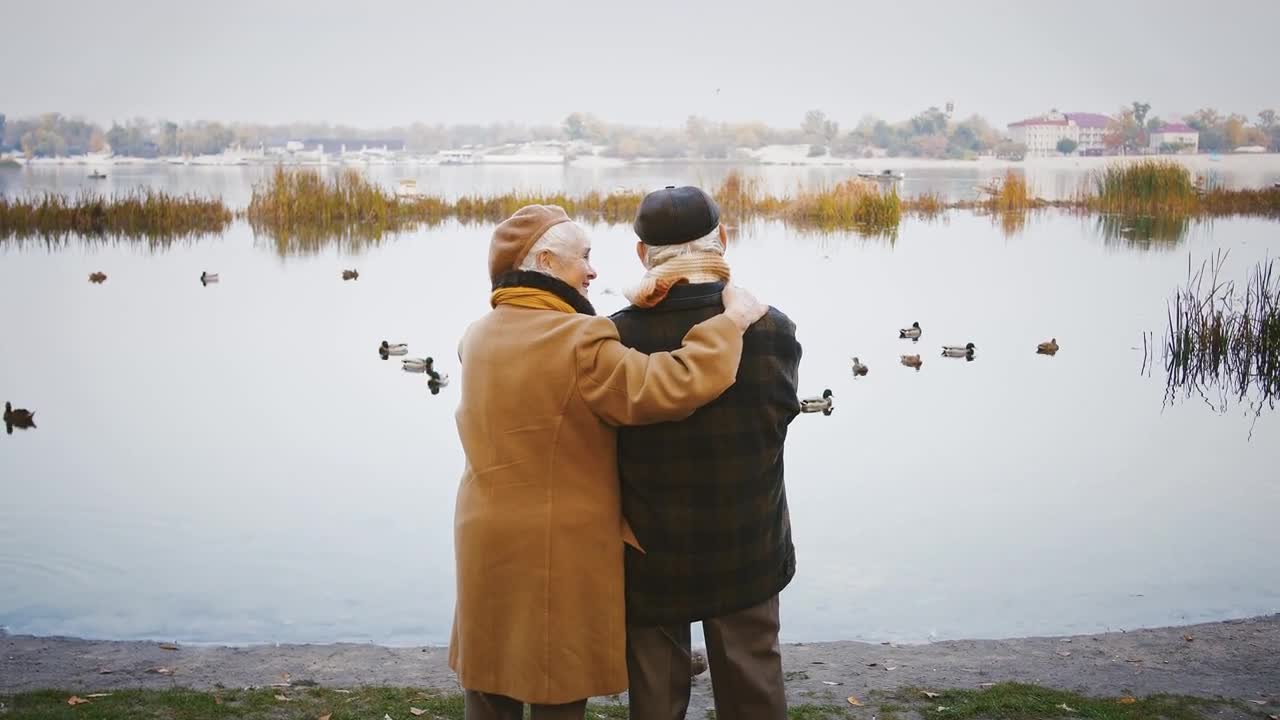 Loving elderly pair hugging and talking, standing on bank of a river with ducks