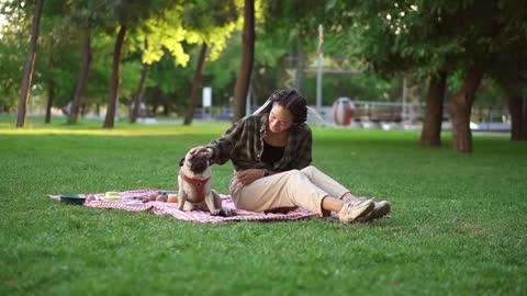 Girl sitting on plaid on lawn in a park while dog sitting in front of her, she pets him