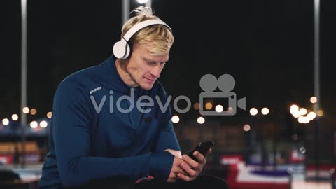 Smiling Sportsman Sitting At Park Listening Music With Bluetooth Headphones, Texting On His Mobile P