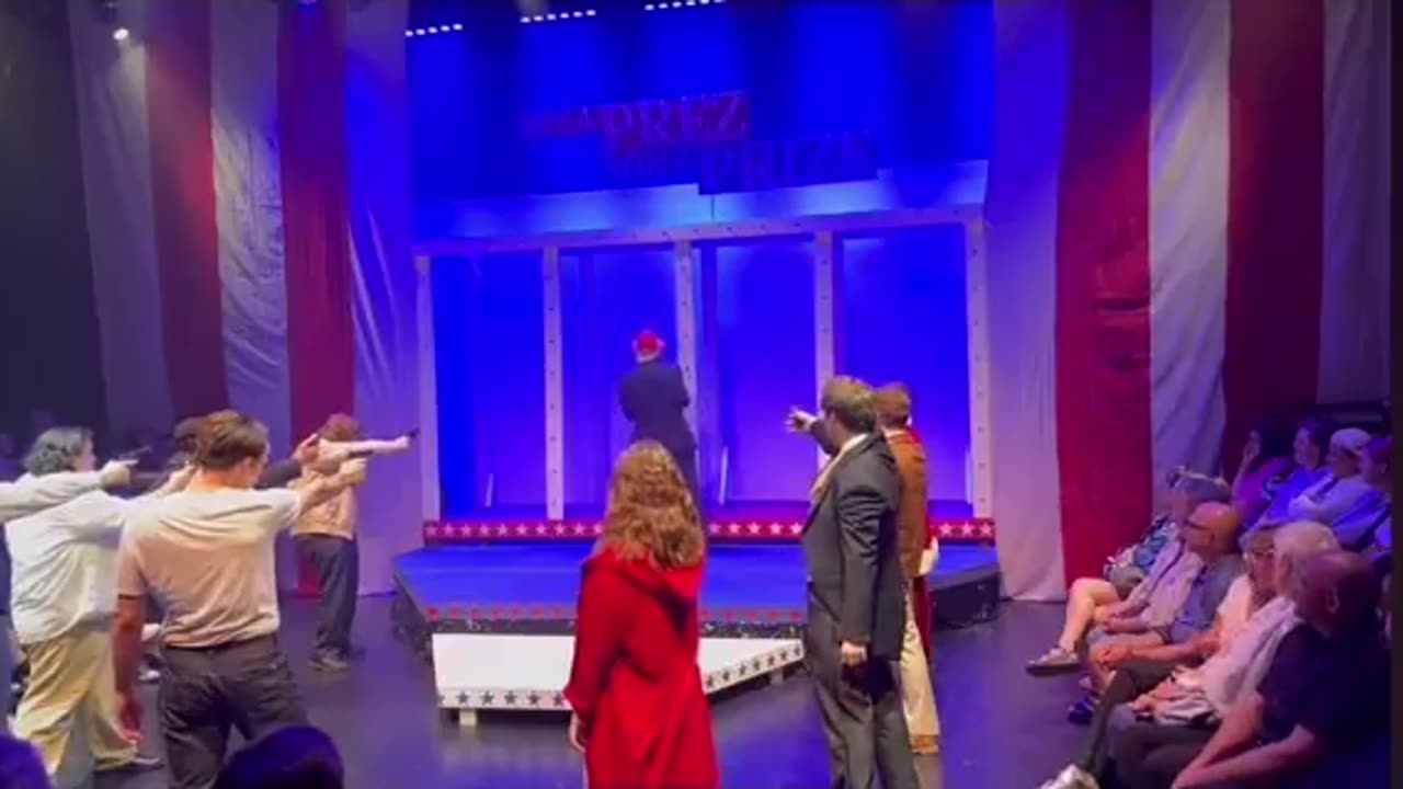 A scene from a play shows a group of people aiming guns at someone dressed up as Trump