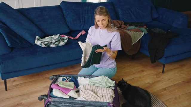 Young cute blonde woman packing clothes into suitcase