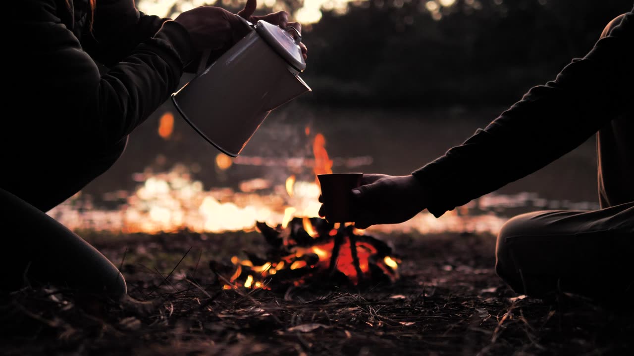 People pouring a warm drink around a campfire