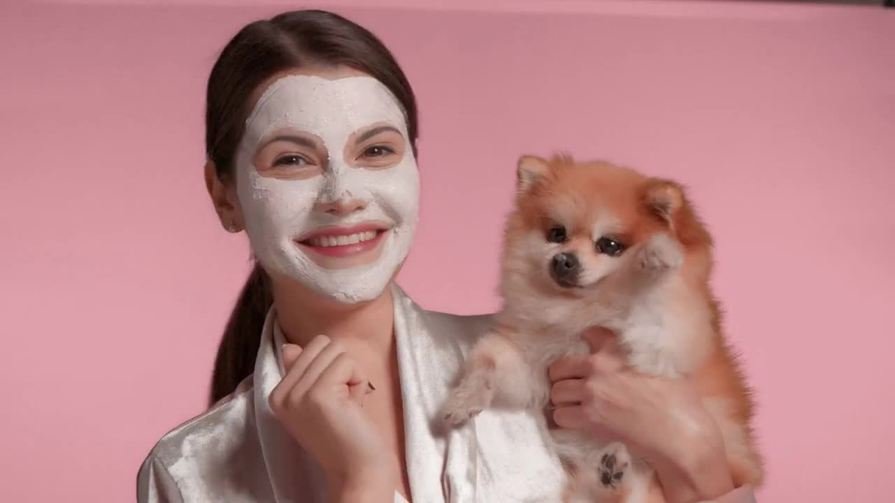 Happy smiling woman in dressing-gown with facial clay mask on and little dog on hands