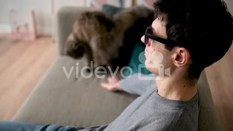 Side View Of A Blind Man In Sunglasses Sitting On The Sofa At Home And Petting His Cat