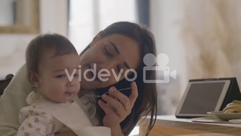 Happy Beautiful Woman Sitting At Kitchen Table And Feeding Her Baby Daughter With Fruit Puree