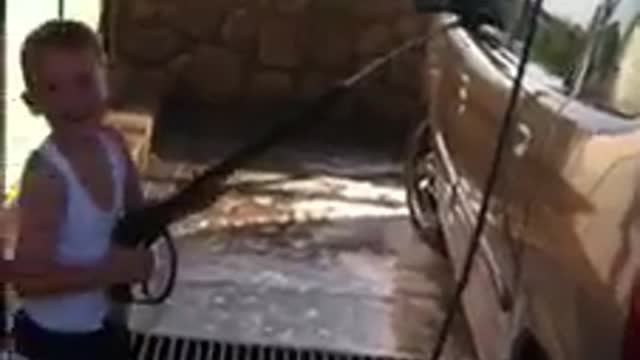 Kid in white shirt trying to wash car gets knocked down by pressure of water