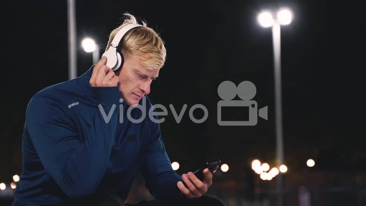 Smiling Sportsman Sitting At Park Listening Music With Bluetooth Headphones And Texting On His Mobil