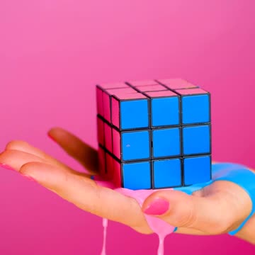 Hand holding a rubik cube that seems to melt on a pink background