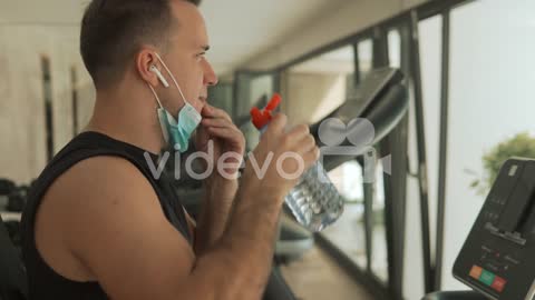 Young Strong Man With Face Mask Uses An Exercise Machine And Drinks Water In The Gym