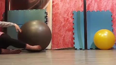 Girl on back on top of grey yoga ball rolls off onto wood floor