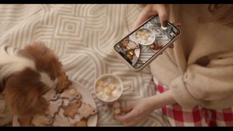 A young girl with her dog sits on a bed with a dish of gingerbread and a cup of cocoa