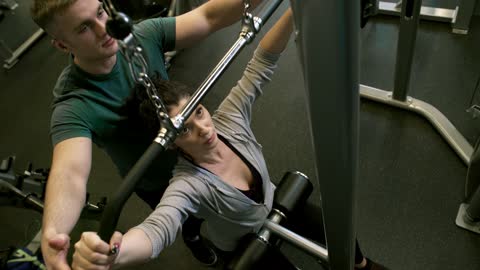 A Man Assisting A Woman In Using The Flat Pulldown Weights Equipment In A Gym