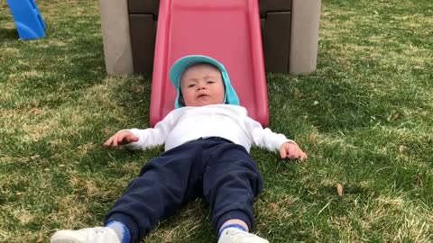 Little girl pushes little boy wearing blue hat down pink slide onto grass