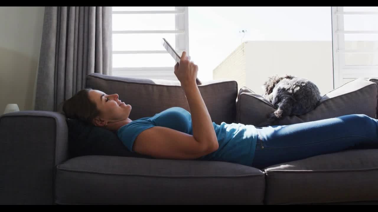 Caucasian woman lying on couch with dog using digital tablet