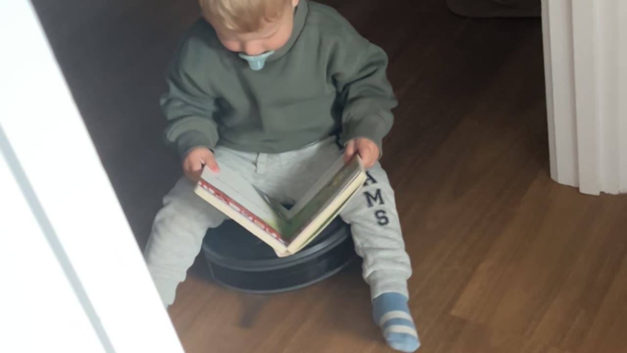 Toddler Reads a Book While Riding Robot Vacuum