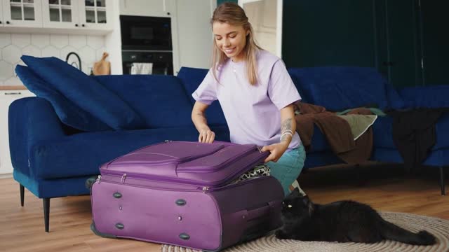 Young positive blonde woman packing clothes into suitcase and closing