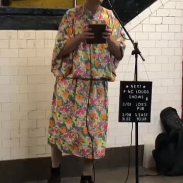 Man dressed up in makeup, wig, and large floral dress performs and sings in a subway station