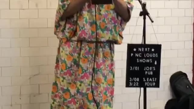 Man dressed up in makeup, wig, and large floral dress performs and sings in a subway station