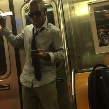 A man in a collard shirt and glasses dances on a subway train