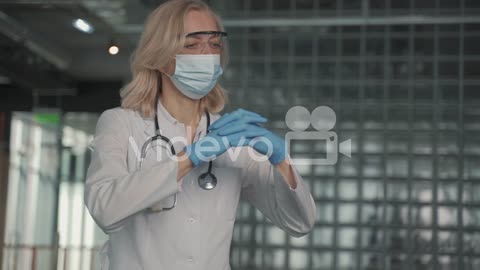 A Blonde, Middle Aged Female Doctor In A Facemask Puts On Surgical Gloves 1