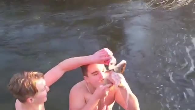 Two guys in river one drinks beer through fish