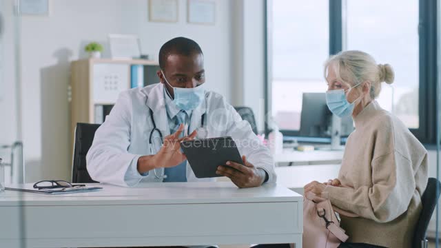 African American Doctor in Protective Mask is Reading Medical History of Senior