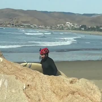 Guy wearing red helmet walking on beach with surfboard