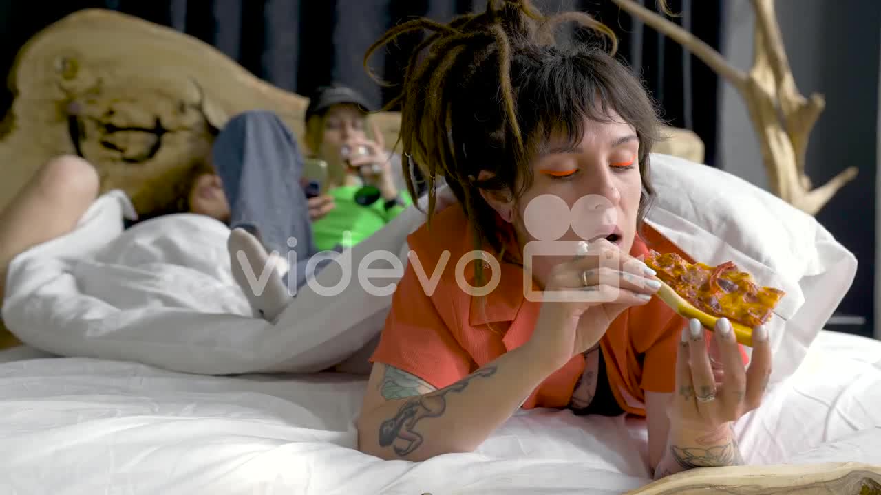 Young Woman With Piercings And Tattoos Eating Pizza While Her Friends Are Behind Drinking Wine And R