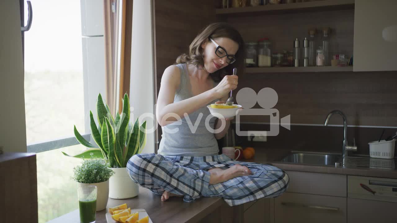 Attractive woman sitting on table and eating breakfast-Young beautiful female in pajamas and eyeglas