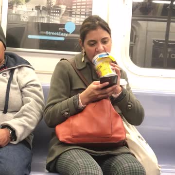 Woman uses her tongue to scoop cereal out of packaged cereal bowl on subway train