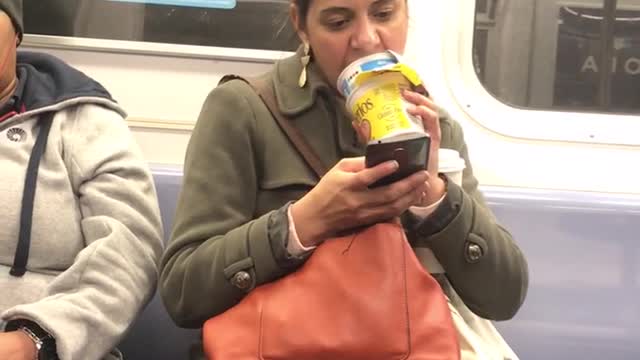 Woman uses her tongue to scoop cereal out of packaged cereal bowl on subway train