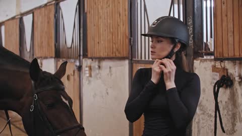 A horsewoman puts on a plastic helmet standing near the horse