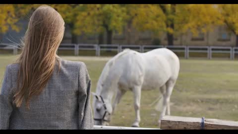 Back view of a brunette Caucasian woman looking at the white horse grazing in the corral