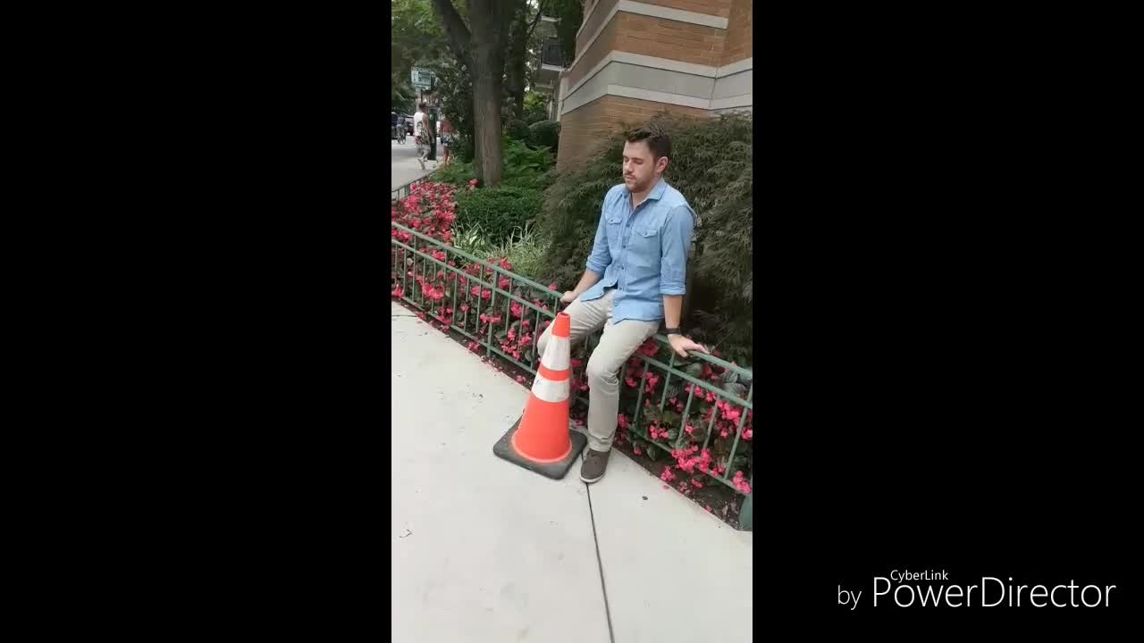 Guy in blue button up and khakis crosses the street and back with orange traffic cone