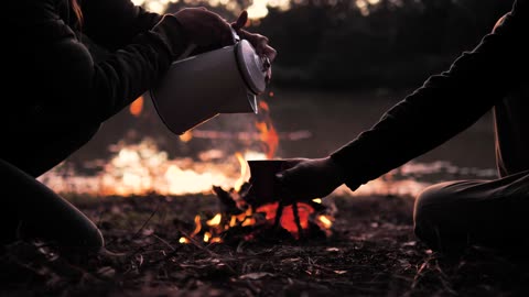 People Pouring A Warm Drink Around A Campfire