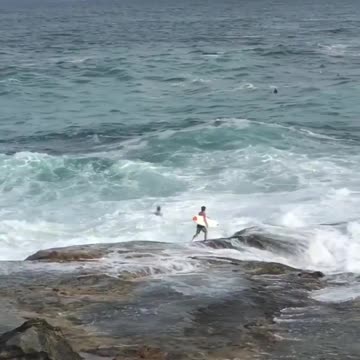 Guy tries to jump into water off rocks at beach but gets pushed back by waves every time