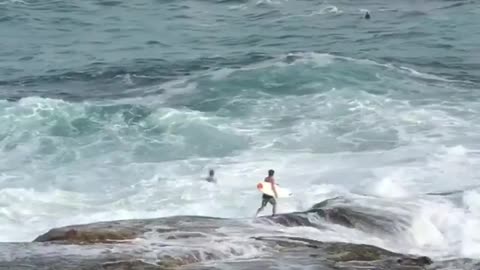 Guy tries to jump into water off rocks at beach but gets pushed back by waves every time