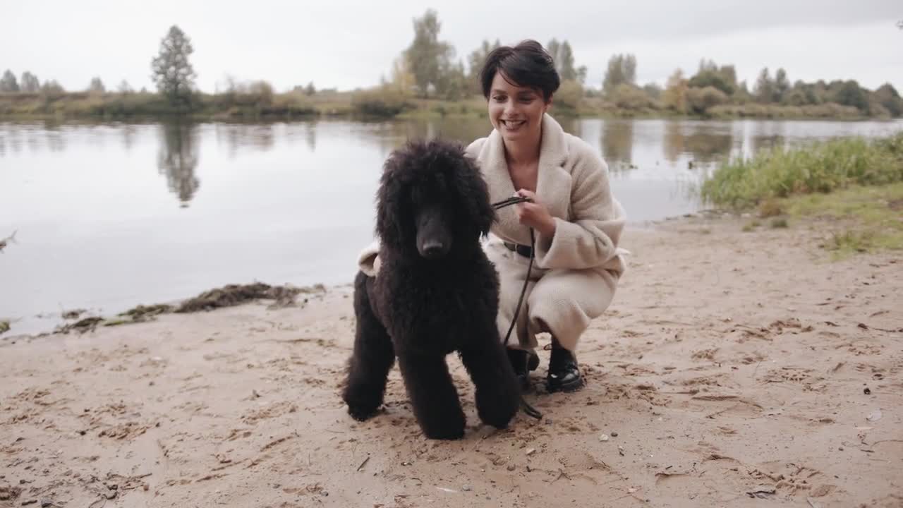 A smiling young woman in a stylish white coat is squatting on the river bank