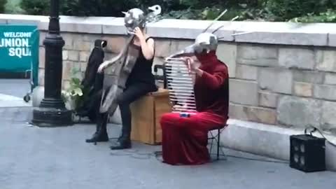 Man on sidewalk floor touching woman's breasts, musicians with metal helmets playing instruments