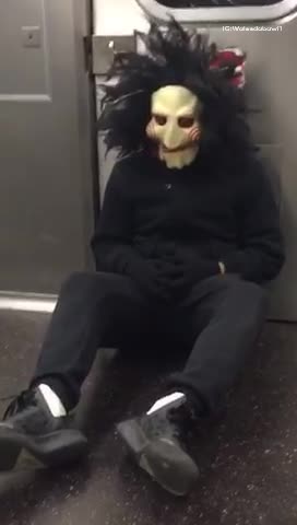 Person dressed as jig saw sitting on subway train floor near door