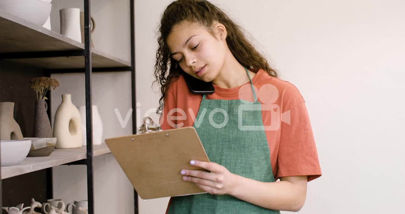 Young Female Clerk Having A Call And Writing On A Clipboard In The Pottery Shop 1