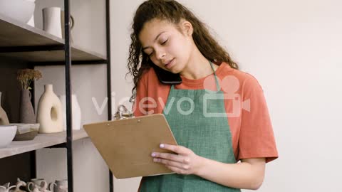 Young Female Clerk Having A Call And Writing On A Clipboard In The Pottery Shop 1