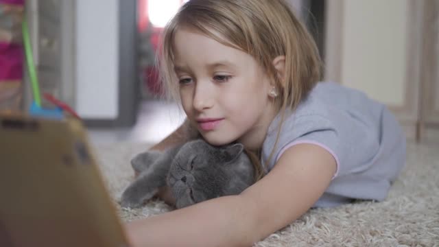 Cute little girl holding digital tablet and hugging your favorite cat lying on the floor