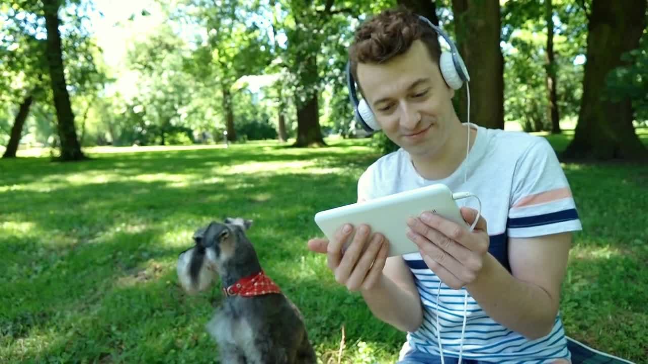 Boy watching something funny on tablet and sitting in the park with his dog, ste