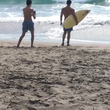 Guy in blue swim trunks takes picture for friends on beach holding yellow surfboard