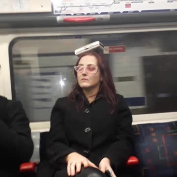Woman with pink glasses balancing black dog container on her head subway