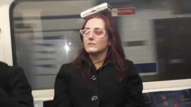 Woman with pink glasses balancing black dog container on her head subway