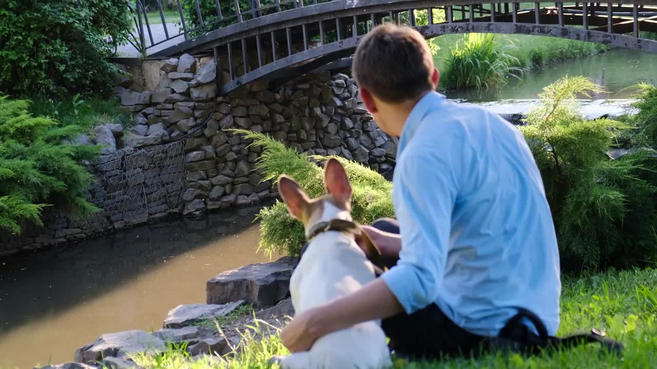 The owner is sitting with the dog by the stream in the park and stroking her