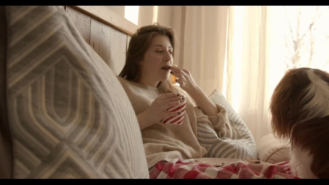 Adorable young woman sitting on the bed leaning on pillows and eating gingerbread